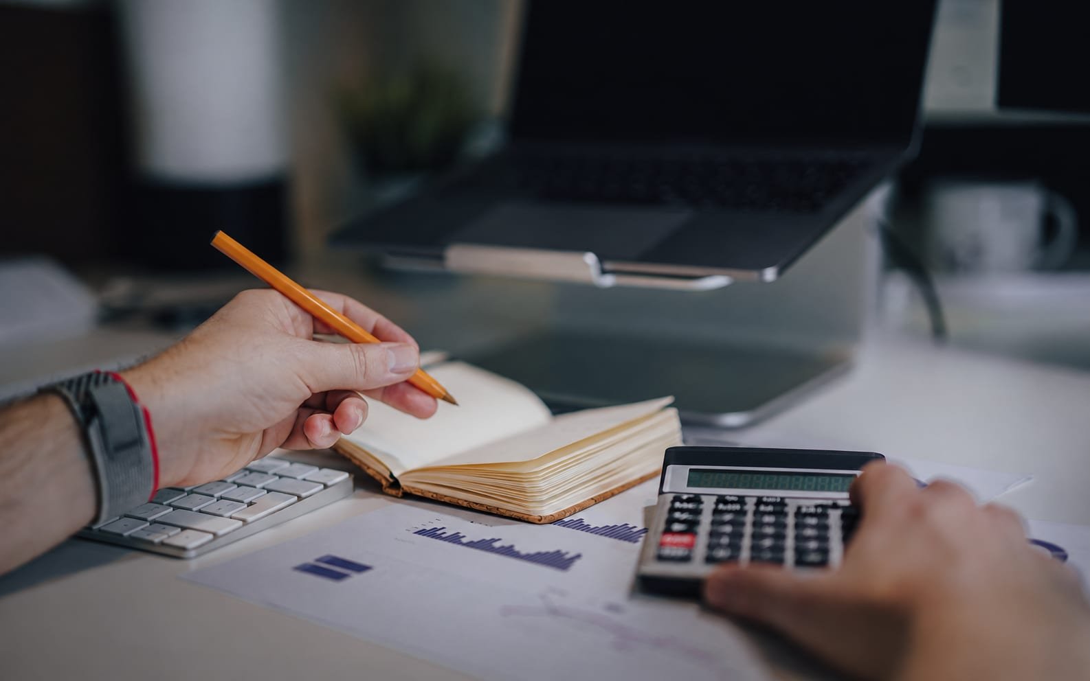 A person reviewing cost figures with a calculator, notebook and a printed bar chart
