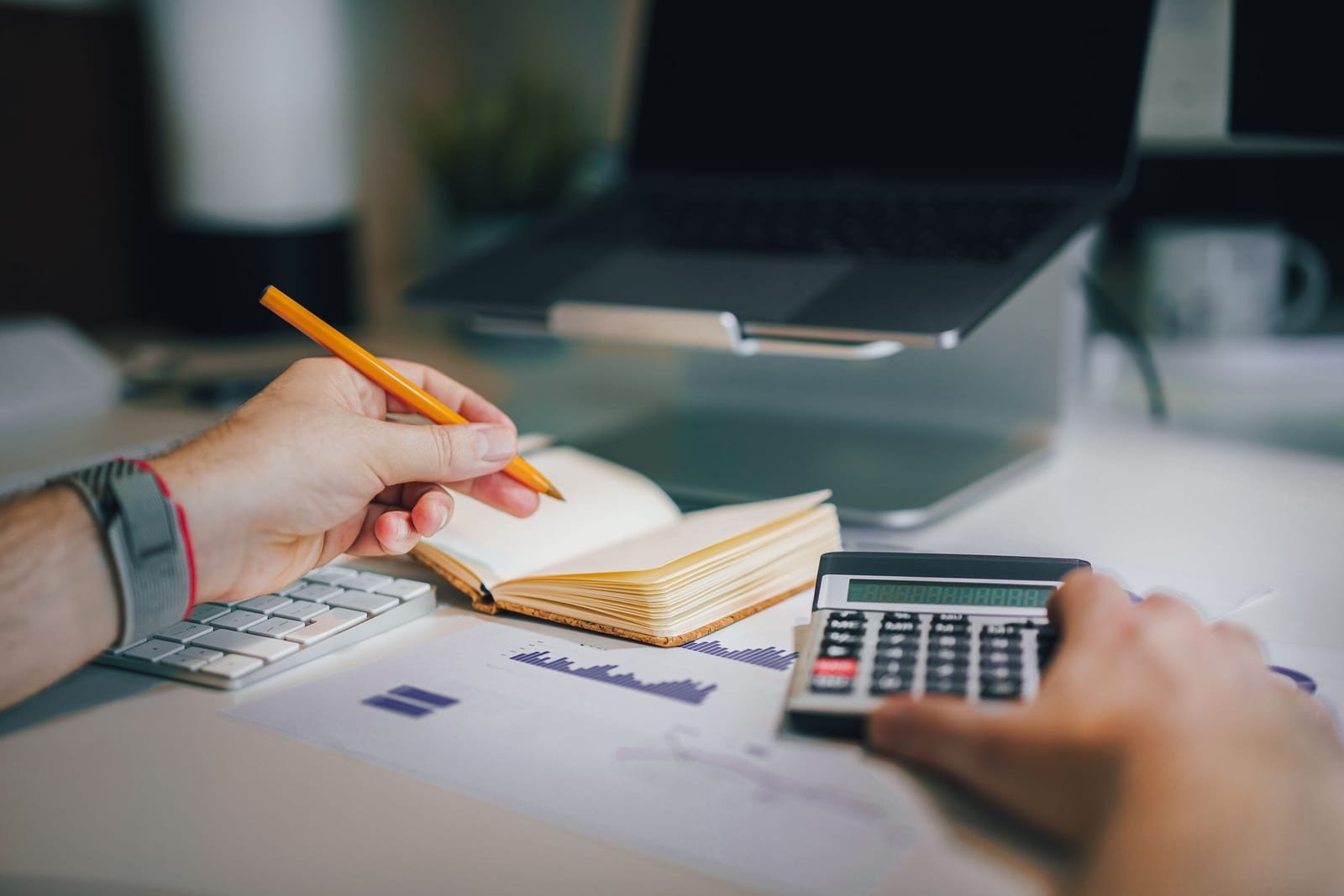 A person calculating advertising budget figures with a calculator, notebook and a printed bar chart